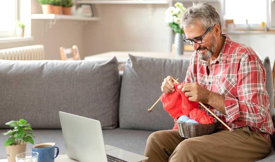 A man knitting on the couch in front of an open laptop