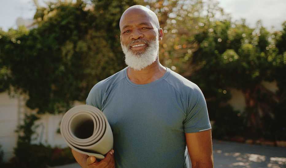 A smiling man holding a yoga mat under his arm outside