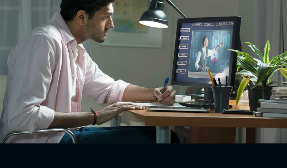 A man writing at a desk as he watches something on a computer