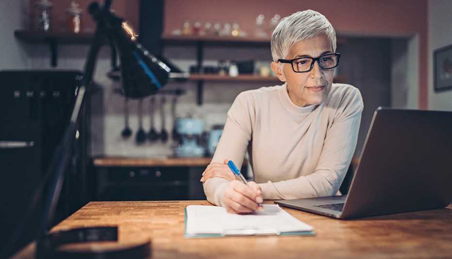 A woman writing at a table at home while looking at a laptop