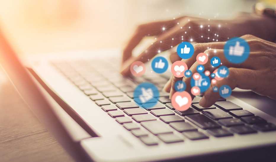 A person is typing on a laptop with Facebook like and love reactions on his hands