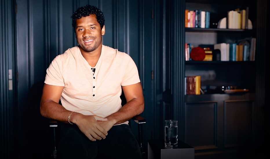 A man, Russell Wilson, sitting and smiling during his presentation on the TED MainStage