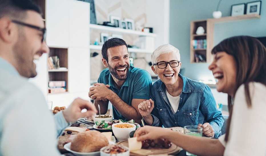 Two men and two women laughing together during a meal at a table in a home