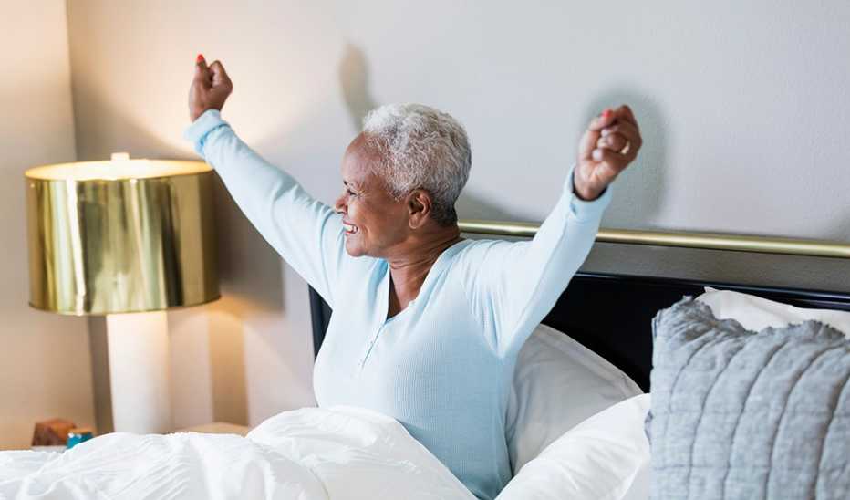 A smiling woman waking up from bed sitting up with her arms stretched